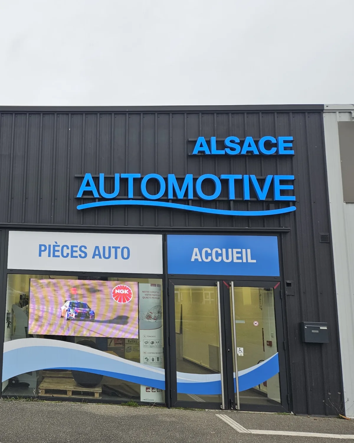 Façade du magasin Alsace Automotive : enseigne lumineuse bleue sur bardage sombre, bandeaux « Pièces auto » et « Accueil »